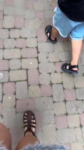 The video shows a person walking on a paved walkway with reddish-brown and tan rectangular pavers. In the foreground, the person's feet are visible, wearing sandals. Further down the walkway, another person, possibly a child, is also walking and wearing sandals and denim shorts. The walkway appears to be in an outdoor setting, possibly a commercial area, with shops and seating areas visible on the right side. Trees and greenery are present on the left side. The camera is moving forward, following the person's walk.