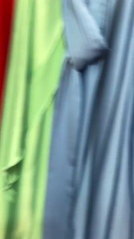 The video shows a close-up, blurred view of a rack of colorful garments, likely clothing. The colors visible include blue, red, yellow, and green. The fabric appears to be flowing or draped, suggesting it might be dresses, skirts, or scarves. The lighting is bright, and the background is mostly indistinct, with hints of white and possibly other clothing items. The camera is moving, creating motion blur that emphasizes the texture and movement of the fabric. There are no people visible in the video.