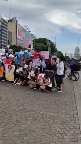 A crowd of people are gathered in an outdoor public area with a large display screen showing Coca-Cola branding. The people are holding up signs and pictures, with some signs clearly showing the name "ENHYPEN." Many of the people are kneeling or sitting on the ground, creating a dense and organized gathering. The background shows buildings and a modern cityscape, with a cloudy sky overhead. One person stands slightly away on a bicycle. The camera pans slightly to the left to capture more of the group and the surrounding cityscape. The people are cheering and chanting.