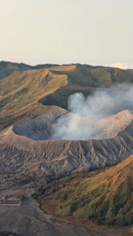 This video shows an aerial view of a volcano crater with smoke rising from its center. The crater is surrounded by hills covered in vegetation. The sky is clear and the lighting suggests it is either sunrise or sunset. The overall landscape is rugged and mountainous. There are no visible people or man-made objects other than what appears to be a small building or structure in the lower left corner of the frame.