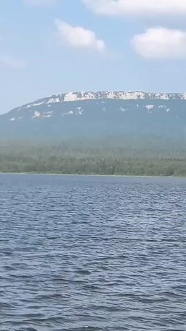 A wide shot shows a calm lake with choppy water in the foreground. Behind the lake, a line of evergreen trees forms the middle ground. A large, flat-topped mountain with patches of snow is visible in the background, under a bright blue sky with scattered white clouds. The video appears to be taken from a boat or shoreline looking out over the water. There are no people or animals visible.
