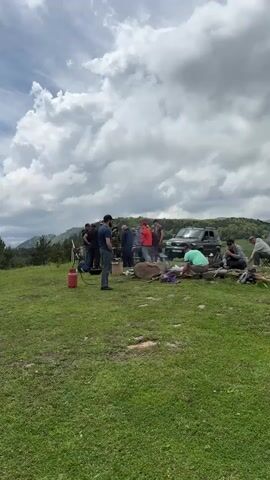 A group of men are gathered in a grassy, hilly area. They appear to be having a picnic or barbecue, with several vehicles parked nearby. One man is tending to a fire, while others are seated or standing around. The sky is cloudy, and the landscape suggests a rural or mountainous region. The video pans across the scene, showing different individuals and activities within the gathering.