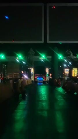 The video shows a nighttime view of a toll plaza. Green lights are illuminated above multiple lanes, indicating they are open. Vehicles appear to be passing through the toll booths, with one truck clearly visible in the center lane, its lights reflecting on the wet ground. The ground is dark and reflects the green and colorful lights from the vehicles and toll plaza structures. Yellow and red traffic barriers are visible on the sides of the lanes. The overall atmosphere is dark with artificial lighting creating a vibrant, reflected glow.
