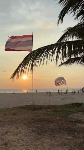 The video shows a beach scene at sunset. The Thai flag is visible on the left. The sun is setting over the ocean in the background. Several people are playing volleyball on the beach. A parachute lies on the beach behind the volleyball players. Palm trees frame the scene. The sky is a mix of orange and yellow hues. Some litter is visible on the sand in the foreground.