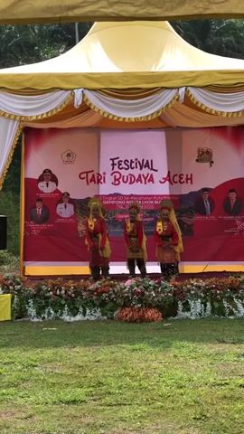The video shows three young girls in traditional Acehnese dance costumes performing on an outdoor stage. The stage is decorated with flowers and has a red backdrop with the words "FESTIVAL TARI BUDAYA ACEH" prominently displayed. The girls, adorned with colorful headpieces and intricate clothing, are moving in unison, their hands and arms in expressive poses. Behind them, portraits of officials are visible on the backdrop. The setting appears to be a public event, possibly a cultural festival, with a grassy area in front of the stage. The lighting suggests it is daytime, and the presence of trees in the background indicates an outdoor environment. The overall atmosphere is festive and cultural.