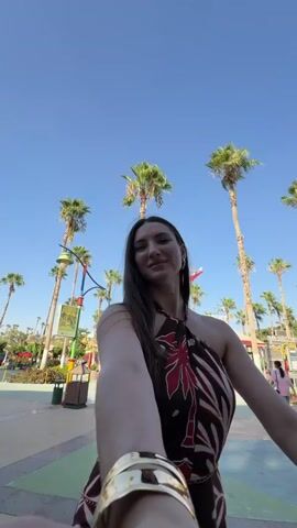 A young woman with long dark hair, wearing a brown and red patterned halter dress and a gold bracelet, is filming a selfie video outdoors. She is smiling and looking at the camera. Behind her, there are several tall palm trees, a blue sky, and what appears to be an amusement park or a similar recreational area with structures and other people in the background. The camera is positioned low and angled upwards, giving a sense of looking up at the woman and the surroundings.