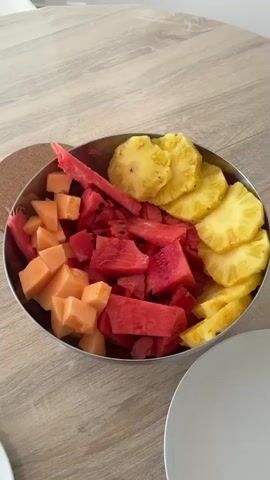The video shows a bowl of fruit salad with cantaloupe, watermelon, and pineapple on a wooden table. There are two white plates on the table, one of which has pasta with red sauce and cheese on it, along with a fork.