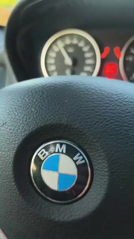 The video begins with a close-up of a car's dashboard and steering wheel, specifically showing the BMW logo. The camera then pans out to show the view from inside the car as it drives on a highway during what appears to be sunset. The road is visible, with other cars ahead and trees lining the sides. The sun is low in the sky, casting a warm glow. The driver's hands are on the steering wheel, suggesting they are actively controlling the vehicle. The speedometer and other dashboard indicators are partially visible, with some red warning lights illuminated.
