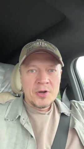 The video features a man inside a car, singing along to music. He is wearing a green baseball cap and a green jacket over a tan shirt. The car's interior is visible, showing beige seats and a black dashboard. The man is looking forward, occasionally glancing at the camera as he sings. The background outside the car window appears blurry, suggesting the car is in motion.