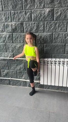 A young girl in a neon yellow crop top and black leggings with black socks performs a dance move in front of a brick wall. She is holding onto a horizontal bar with her left hand and has her right leg extended straight up in the air, her left leg is bent at the knee. A white radiator is visible to the right of the girl.