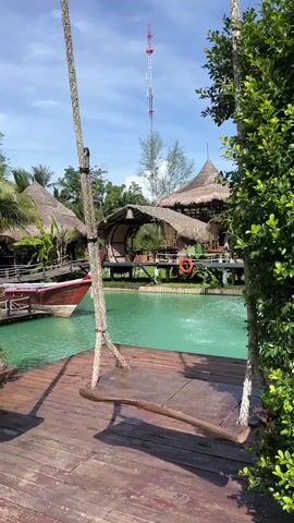 The media file captures a tranquil scene in Tambon Pa Kae Bo Hin, Thailand, likely during the daytime under a clear blue sky with scattered clouds. The primary subject is a rustic wooden swing suspended by thick ropes from two trees or posts, positioned on a weathered wooden deck. The swing faces a vibrant turquoise body of water, possibly a lagoon or a canal.

In the background, traditional Thai architecture is evident with several thatched-roof huts and structures built over the water, connected by wooden walkways. A longtail boat, a distinctive Thai vessel, is docked to the left, its red and white bow adorned with a red and white ribbon. A life preserver ring is visible on one of the structures. Further in the distance, a tall communication tower pierces the sky. Lush green foliage frames the right side of the image, adding to the tropical ambiance.

The scene suggests a peaceful, idyllic setting, perhaps a resort or a cultural attraction. The water is calm, with gentle ripples indicating some movement, possibly from a small fountain or the boat. The sunlight casts shadows on the wooden deck, indicating it's a bright day. There are no people visible in the frame, contributing to the serene and undisturbed atmosphere. The overall emotion conveyed is one of relaxation and natural beauty. No visible text is present in the media file.