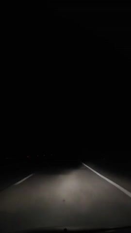A low-angle, interior shot from a vehicle moving forward on a dark highway at night. The vehicle's headlights illuminate the gray asphalt and white dashed lines separating lanes. The distant horizon is barely visible with a few small red lights in the far distance.