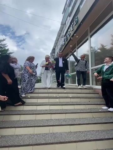 The video captures a celebratory scene on a set of outdoor stairs in front of a building. A couple, the man in a black suit and the woman in a white pantsuit, are descending the stairs, holding a bouquet of flowers. They are being showered with rose petals by a group of people standing on and around the stairs. The man is holding a small basket adorned with ribbons and flowers. The overall atmosphere appears to be joyful and festive.