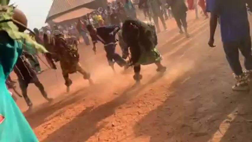 The video shows a group of people, some wearing masks and traditional attire, in a dusty outdoor area. The scene is chaotic with people running and kicking up dust. One person is lying on the ground. The camera is hand-held and shaky, focusing on the action in the center of the scene while a person holding a branch stands in the foreground. There are buildings and many spectators in the background.