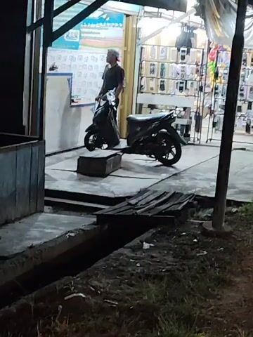 A lone individual stands outside a shop at night. The shop appears to be a retail establishment, possibly selling phone accessories, judging by the displays visible through the open doorway. Two motorcycles are parked near the entrance. The area surrounding the shop is unlit and includes a drainage ditch. The video is static, showing no movement other than the person's slight shifts in posture.
