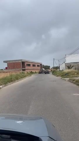 The video shows a paved road on a cloudy day. A black car drives away in the distance. Parked on the side of the road is a red tractor with a large water tank attached to it. There is a building under construction to the left of the road and an existing white building to the right. There are some weeds and greenery near the buildings. Power lines are visible above the right side of the road. The footage is taken from inside a moving vehicle.