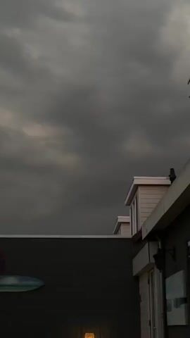 The video shows a dark, cloudy sky above buildings. The camera is positioned looking upwards. On the right side of the frame, there is a building with white trim and windows. There is a darker colored building in the foreground on the left with a grey wall and a decorative surfboard-like object mounted on it. The lighting suggests it is dusk or an overcast day.