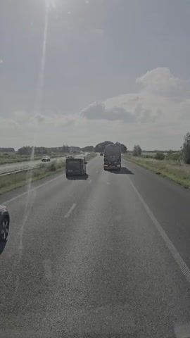 This is a first-person perspective video, likely captured from inside a vehicle, showing a highway scene in Steenbergen, Netherlands. The sun is bright, suggesting it is daytime, with some scattered clouds in the sky, indicating fair weather. The road is asphalt and appears to be a multi-lane highway.

Several vehicles are visible on the highway. In the foreground, on the left lane, a dark-colored SUV is driving away from the camera. Further ahead in the same lane, a white car is also visible. In the right lane, there is a large truck with a trailer, and behind it, a smaller van. In the distance, on the left side of the highway, another car can be seen. The surrounding landscape is flat and rural, with fields and trees lining the sides of the road. There is a body of water to the left of the highway.

The activity taking place is vehicular traffic on a highway. The overall mood conveyed is one of travel and movement. The image is bright and clear, with the sun's glare occasionally affecting visibility. No text is visible in the video.