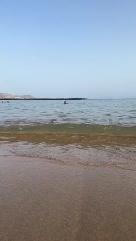 The video opens with a close-up shot of a wave breaking on a sandy beach under a clear blue sky. As the camera pans out, it reveals a family, including two adults and two children, walking into the shallow water. The father is holding the hand of one of the children, and they are all facing away from the camera. The scene then transitions to a wider view of the beach, showing numerous people swimming, walking in the water, and sunbathing. Palm trees and resort buildings line the background, with hills visible in the distance. The water is calm, with gentle waves lapping at the shore. The overall impression is a pleasant day at a crowded beach resort.
