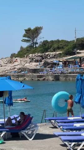 A vertical, medium shot captures a sunny day at a rocky beach with calm, blue water. In the foreground, a woman reclines on a blue and red lounge chair under a blue umbrella, looking at her phone. To her right, a boy stands on a concrete walkway, holding a large, deflated blue inflatable ring. Further back, a person lies on a paddleboard in the water. The background shows more lounge chairs and umbrellas on a rocky shore, with trees and vegetation on a hill overlooking the area. People can be seen scattered on the beach and near some structures. The sky is clear and blue.