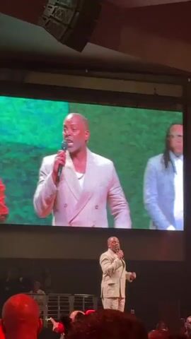 A video shows a live performance taking place in a large venue. In the foreground, an audience can be seen from behind, with heads and shoulders visible. They are facing a stage. On the stage, a large screen displays a live feed of performers. The performers on screen are a man and a woman dressed in formal attire. The man is wearing a white suit and holding a microphone, appearing to be singing or speaking energetically. The woman, to his left, is wearing a black and red dress and also appears to be performing. Below the screen, on the main stage, another man in a light-colored suit is also holding a microphone and performing, gesturing with his hands. He is further from the camera than the performers on the screen. The overall environment appears to be a concert or a religious service, with a focused and engaged audience.