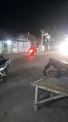 The video depicts a street scene at night, likely in a tropical or subtropical region. The lighting suggests artificial illumination from various sources, including streetlights and shopfronts. In the foreground, a bamboo table is visible, with what appears to be a motorcycle parked nearby. The background shows buildings, some with lights on, and other vehicles.

At approximately 00:03, a person wearing a patterned dress and a headscarf is visible from the back, standing near a stall or shop. Another person, a man wearing a blue shirt and a sarong, is seen in the next frame at 00:04, also facing away from the camera, standing near the same stall. The overall atmosphere seems to be that of a bustling market or roadside area operating into the evening. The video is somewhat shaky, indicating it was likely filmed handheld.