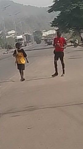 Two individuals wearing dark shorts and athletic tops (one red and one yellow) are seen on a road. They are walking quickly away from the camera, seemingly carrying objects on their backs. The surrounding environment includes trees, buildings, utility poles, and possible vehicles. The location appears to be an urban area with a somewhat hazy background suggesting the potential presence of smog or dust in the atmosphere.