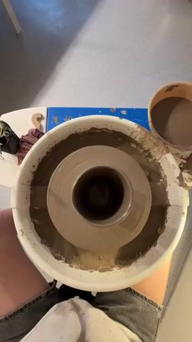 This is a top-down, close-up shot of a pottery wheel with clay on it. The pottery wheel is white and has a circular basin around the edge, filled with brown clay and water. In the center of the wheel, a lump of clay is spinning and being shaped into a pot. The camera angle is directly above the wheel, looking down. Parts of a person's legs and torso are visible at the bottom of the frame, indicating they are operating the wheel. To the right of the pottery wheel, there is a bucket filled with brown clay and water. The background appears to be a light-colored floor, possibly concrete, and a white surface to the left, which might be a table. The lighting is natural and appears to be coming from above.