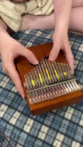 A person's hands are shown playing a kalimba. The kalimba is made of wood and metal, with metal tines that produce sound when plucked. The kalimba has yellow markings on some of the tines, which may indicate specific notes. The person is sitting on a blue and white checkered blanket. The background is out of focus, but appears to be a person wearing light-colored shorts. The video captures the action of playing the kalimba.