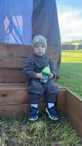 A small child is sitting on a wood box on a lawn. The child is wearing a grey hat, a grey onesie, white socks, and blue sneakers. The child is holding a blue water bottle with a green top. Behind the child is a brown and blue tent-like structure. In the background, there is green grass, trees, a few houses, and a bright blue sky. Someone out of frame says, "Синий трактор едет к нам" which translates to "Blue tractor is coming to us." The child responds with indiscernible words.