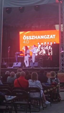 The video shows a live music performance taking place on a stage under a canopy. A large LED screen behind the performers displays the text "ÖSSZHANGZAT 4.0" in orange and white, along with images of musicians. Two individuals are on stage, center-left. The person on the left, with long hair, is wearing a light-colored patterned shirt and light-colored pants. They appear to be playing a keyboard or synthesizer. The person on the right is wearing a light blue button-up shirt, a grey vest, and light-colored pants, and is holding a microphone. They are singing or speaking into the microphone. Other band members are visible on the stage behind them, including a person playing a saxophone. In the foreground, an audience is seated on chairs, facing the stage. The chairs have "GALERIA" printed on them. The lighting is dim, with red and pink lights illuminating the stage from the left.
