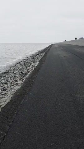 The video shows a grey paved road running alongside a shoreline. The sea, also grey, is to the left of the road. The waves are lapping against a shore reinforced with dark rocks. The sky is overcast and a uniform grey. In the distance, several people are walking along the horizon, appearing as small silhouettes. The camera seems to be at ground level and slightly tilted upwards.