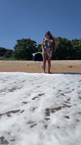 A video shows a woman standing on a sandy beach in Sant Pol de Mar, Spain, under a bright, clear blue sky during the daytime. Her long, light brown hair, moved by the wind, is visible. She is wearing a loose, short-sleeved top or short dress with a vibrant, multi-colored pattern, possibly over white shorts. She stands barefoot at the water's edge, facing the camera, as foamy ocean waves wash over her feet and recede. In the background, a line of green trees borders the beach, and a small, dark beach tent or shelter is visible among them. The sun is high, casting distinct shadows. The woman makes slight hand gestures near her face, possibly adjusting her hair or shielding her eyes from the bright sunlight. The sound of ocean waves is audible.