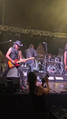 A live band is performing on a stage with bright blue and white lights. In the foreground, a woman with dark hair is filming the performance with a camera. On stage, from left to right, a man in a cowboy hat and a leather vest is playing a red electric guitar. Behind him, another man is playing the drums. A singer with long, dark hair and a leather vest is standing center stage, holding a microphone and singing. To the right of the singer, a bald man in a black vest is playing a guitar. The stage has a raised platform for the drums and is backed by lighting rigs and a backdrop with a logo.