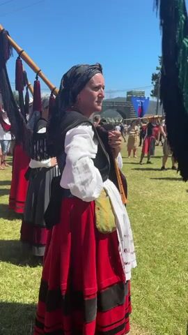 A woman in traditional clothing, including a red skirt with black trim, a white blouse, a dark vest, and a headscarf, stands in a grassy field holding a wooden wind instrument. In the background, a group of people in beige tunics and some in red dresses and black vests are assembled. The people in the background are holding flags and what appear to be ceremonial staffs. The sky is clear and blue, and there are trees and banners visible in the distance, suggesting an outdoor event or festival. The focus is on the woman in the foreground, while the background provides context for a gathering.