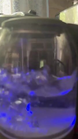 The video shows a close-up of a glass electric kettle filled with water that is boiling. The water is agitated and appears frothy with steam rising. The kettle has blue LED lights at its base, illuminating the boiling water. Visible markings on the kettle indicate a maximum fill line ("MAX") and volume markers for "1.0L" and "0.5L MIN". The brand name "Mampēna" is visible at the base of the kettle. The background is blurry but suggests an indoor setting, possibly a kitchen, with some objects and possibly a window with natural light. The overall impression is a common household activity of boiling water, highlighted by the dynamic visual of the boiling water and the blue light. There are no people visible in the frame. The time of day and weather are not discernible. The location context provided is Zubov, Russia, but there are no specific visual cues within the video to confirm this location.