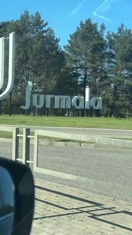 The video shows a car driving past a welcome sign for Jūrmala, Latvia. The sign is large, white, and modern in design, with several tall, curved elements next to the name "Jūrmala" in large, sans-serif letters. The sign is on a green lawn with trees in the background. The camera is inside a moving car. There is a blue directional sign with a white arrow next to the welcome sign.  Beside the road, three electrical boxes stand on the grass. The day is sunny.