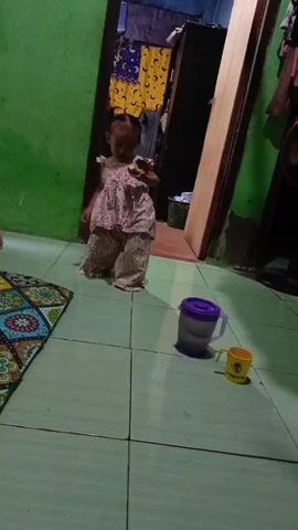 A toddler in a patterned dress and pants stands in a room with green walls and a tiled floor. Next to the toddler are a purple lidded pitcher and a yellow cup. A young boy in a patterned shirt and blue shorts enters the frame, holding a flashlight. The boy then crouches down near the toddler, and they both interact with the flashlight and what appear to be small objects on the floor.