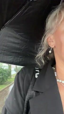 A person with blonde hair wearing a black coat and pearl earrings holds a black umbrella while walking in the rain on a paved path lined with trees. The path is wet and appears to have puddles. The person is looking off to the side, and the video is shaky. A glimpse of a white van and a yellow traffic sign is visible in the background.