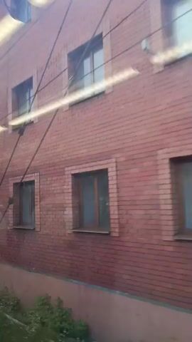 The video shows a view from a moving vehicle looking out a window. The primary focus is on the exterior of a brick building with several windows. There are also some signs of graffiti on the building. In the background, there is a concrete wall, some greenery, and the sky with clouds. A red building with the word "Motel" is visible in the distance towards the end of the clip. The overall impression is that of passing by an urban or suburban area.