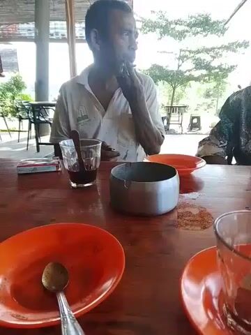 A video shows two men sitting at a wooden table outdoors. The man on the left, wearing a white shirt, is seen with his hand to his mouth, possibly eating or smoking. In front of him is a glass with a dark liquid and a cigarette pack. To his right is a metal ashtray. The man on the right, wearing a patterned shirt and a dark cap, is seen with his hands in front of him. In front of him is a glass of water and an orange saucer with a glass of red liquid. Two orange plates with spoons are also visible on the table. The background suggests an outdoor setting with trees and some structures, possibly a restaurant or cafe. The lighting is bright, indicating daytime.