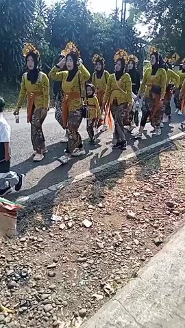 A group of people, primarily women and two young children, are walking down a paved road. They are dressed in traditional attire, consisting of yellow tops, patterned sarongs, and ornate headpieces. The women are wearing hijabs. One of the children is carrying a small white object. The group is walking in the same direction, with the women appearing to lead the procession. The road is lined with trees and vegetation on the left side, and a gravel shoulder on the right. Sunlight is casting shadows on the road, indicating it is daytime. A white motorcycle is visible in the background on the right. The overall scene suggests a parade or a cultural procession.