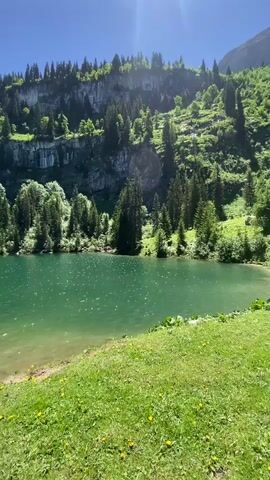 The video opens with a wide shot of a vibrant turquoise lake surrounded by lush green trees and imposing rocky cliffs under a bright blue sky with visible sun glare. The camera pans slightly to reveal a grassy area in the foreground, dotted with yellow wildflowers. A pair of dark hiking boots are prominently placed on the grass. As the video progresses, the camera zooms out slightly and pans left, showing a person standing in the shallow water at the edge of the lake, facing away from the camera and towards the water. The person is wearing a dark t-shirt and shorts. The scene is serene and natural, suggesting an outdoor recreational setting.