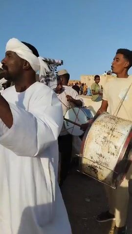 Here's a description of the video:

The video shows a group of people in a festive atmosphere, likely at an outdoor event or celebration. There are at least three men playing drums. One is holding a large, well-worn drum, while another uses sticks to play a smaller drum. A man wearing a white robe and head covering is dancing, raising his hands enthusiastically. Other people are visible in the background, some of whom appear to be clapping along. The setting is dusty, with a clear blue sky overhead. A building is visible in the background with other people standing or sitting in its vicinity.