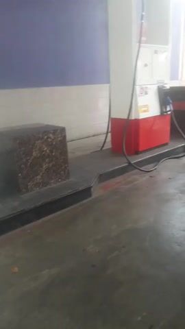 The video shows a gas station interior. A person wearing a white helmet and a gray and white shirt is standing next to a motorcycle. Another person, wearing a white shirt and black pants, is also near the motorcycle, holding a gas nozzle. The motorcycle has a license plate that reads "K 6180 VR 12-25". A gas pump with red and white colors is visible, along with a marble-like block to the left. The ground is concrete and wet in some areas. The overall lighting suggests daytime.