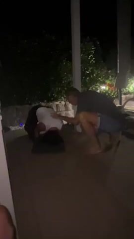 Two people are on a paved patio at night. One person is on their hands and knees facing away from the camera, with their head down. The other person is crouching nearby, facing the person on the ground and touching their back. In the background, there are trees and lights.