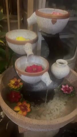 This is a medium shot of an outdoor water fountain. The fountain is made of ceramic and has three tiers of bowls. Each bowl has a spout from which water flows into the bowl below. The bowls are speckled white with gold trim. There are floating flowers in the bowls and the main basin of the fountain. The flowers are red, yellow, and pink. Behind the fountain is a blurred background of green plants and wooden furniture.