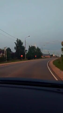 A car is driving down a rural road at dusk. The road curves to the right, and there are power lines and trees on either side. There are also some houses in the distance. The sky is a pale blue and there are a few clouds. The video is shot from inside the car, looking out through the windshield.