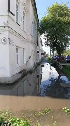 A video captures a side view of a dilapidated building with large windows and peeling paint. The building appears to be under construction or undergoing repairs, with visible cracks in the facade. A large tree stands to the right of the building, its branches casting shadows on the ground. The street in front of the building is flooded with murky water, reflecting the building and the tree. Green grass and weeds grow along the edge of the flooded area. The scene is set on a sunny day, with a clear blue sky visible in the background. There are no people or vehicles visible in the frame.
