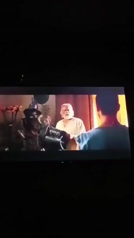 Two individuals are in a dimly lit room, likely indoors, with a large screen displaying a scene. The scene shows an elderly man with a white beard and hair, wearing a light-colored robe, gesturing with his hands. He is facing another person, whose back is to the camera, wearing a blue shirt. To the left of the elderly man, there is a display with various objects, including a mannequin wearing a black hat and a dark robe, and a vase with flowers. The background appears to be a richly decorated interior with curtains. The second person in the scene, whose back is to the camera, is now holding a dark, spherical object with metal embellishments. The lighting in the room is subdued, with the screen being the primary light source for the viewers.